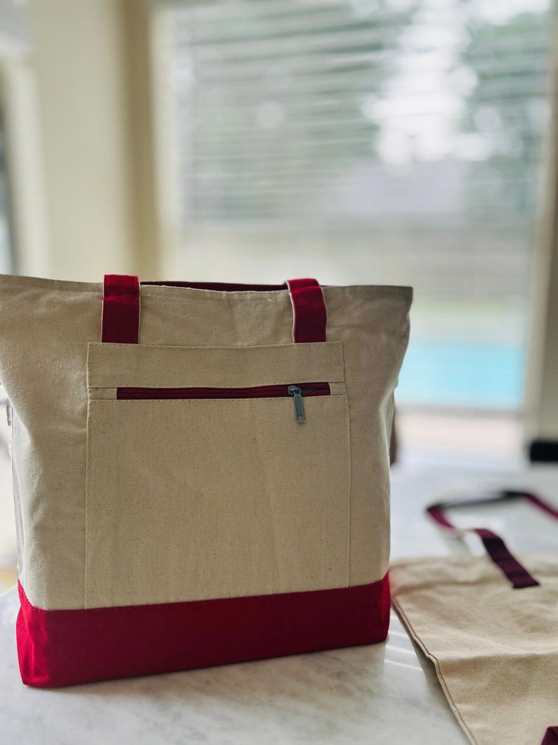 May include: A beige canvas tote bag with a red trim and a zippered pocket. The bag has two red straps.