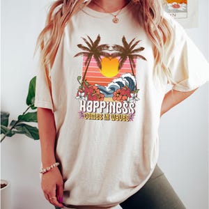 May include: A cream-coloured t-shirt with a tropical design. It features palm trees, a sunset, and ocean waves. The text "Happiness comes in waves" is displayed. The t-shirt has short sleeves and is a relaxed fit.