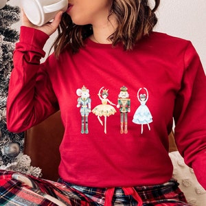 May include: Red long-sleeved shirt with a watercolor illustration of the Nutcracker and his friends. The illustration features a mouse king, a ballerina, the Nutcracker, and a sugar plum fairy.