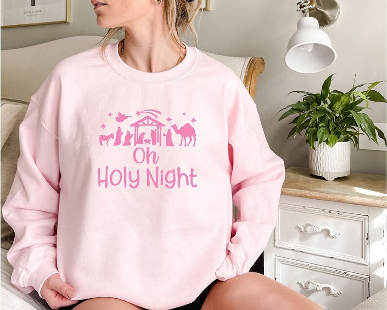 K&ouml;nnte beinhalten: Rosafarbener Pullover mit einem rosafarbenen Motiv einer Weihnachtskrippe und dem Text "Oh Holy Night".