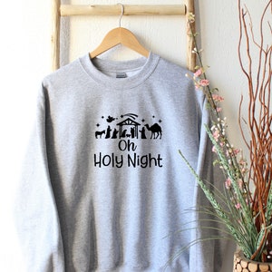 K&ouml;nnte beinhalten: Ein graues Sweatshirt mit einer schwarzen Silhouette einer Weihnachtskrippe und dem Text "Oh Holy Night".