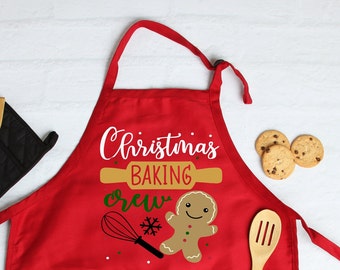 May include: Red Christmas baking crew apron with a gingerbread man, a rolling pin, and a whisk. The text on the apron says "Christmas Baking Crew".