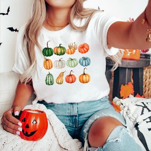 May include: White t-shirt featuring a watercolor design of various pumpkins in shades of green, orange, and white. The pumpkins are arranged in a grid pattern. A person is holding an orange pumpkin-shaped mug.