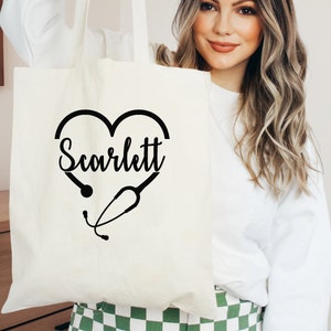 May include: White canvas tote bag with a black heart design featuring a stethoscope and the name "Scarlett" printed inside the heart.