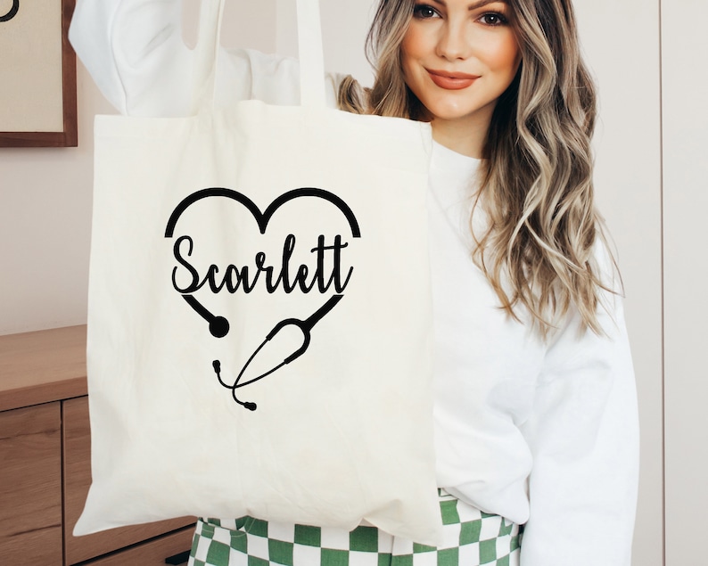 May include: White canvas tote bag with a black heart design featuring a stethoscope and the name "Scarlett" printed inside the heart.
