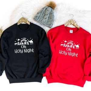 K&ouml;nnte beinhalten: Zwei Weihnachtspullover, einer schwarz und einer rot, mit einer wei&szlig;en Grafik einer Weihnachtskrippe und dem Text "Oh Holy Night".