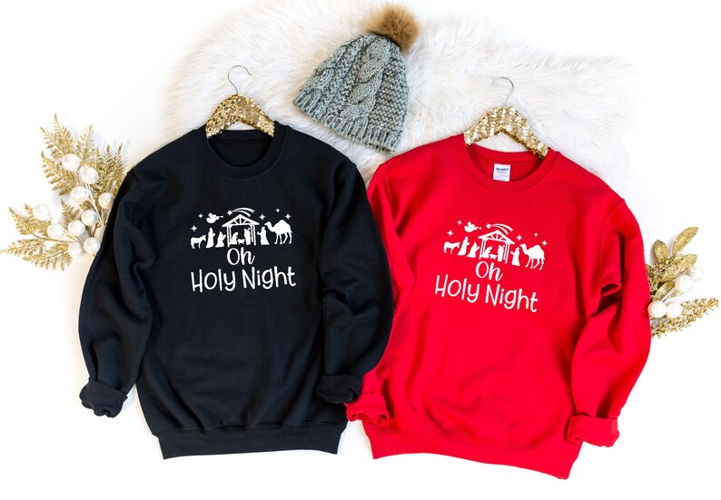 K&ouml;nnte beinhalten: Zwei Weihnachtspullover, einer schwarz und einer rot, mit einer wei&szlig;en Grafik einer Weihnachtskrippe und dem Text "Oh Holy Night".