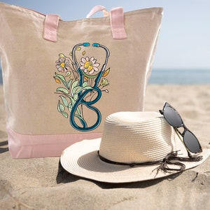 May include: A beige canvas tote bag with pink handles and a blue stethoscope with floral accents. A straw hat with a brown ribbon and black sunglasses are on the sand next to the bag.
