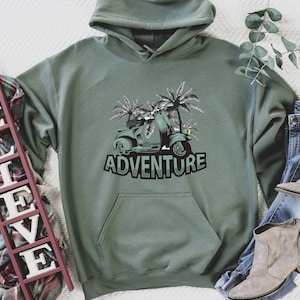 May include: A green hooded sweatshirt with a graphic of a scooter driving through palm trees. The text "ADVENTURE" is printed below the scooter.