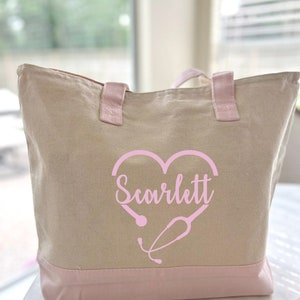 May include: A beige canvas tote bag with pink handles and a pink bottom. The bag has a pink heart with a stethoscope design and the name "Scarlett" written inside the heart.
