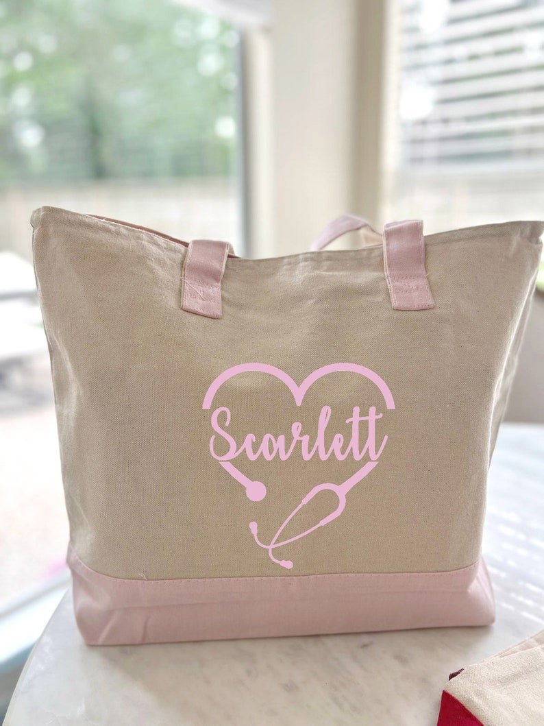 May include: A beige canvas tote bag with pink handles and a pink bottom. The bag has a pink heart with a stethoscope design and the name "Scarlett" written inside the heart.
