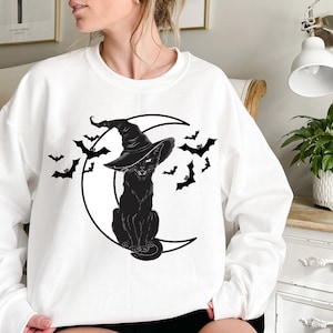 May include: White sweatshirt featuring a black cat wearing a witch hat, sitting on a crescent moon. Black bats are flying around the moon. The design is a Halloween theme.