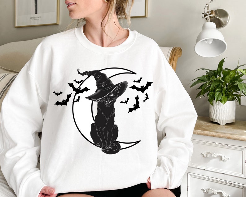 May include: White sweatshirt featuring a black cat wearing a witch hat, sitting on a crescent moon. Black bats are flying around the moon. The design is a Halloween theme.