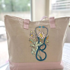 May include: A beige canvas tote bag with pink handles and a pink bottom. The bag has a floral design with a stethoscope in the center.