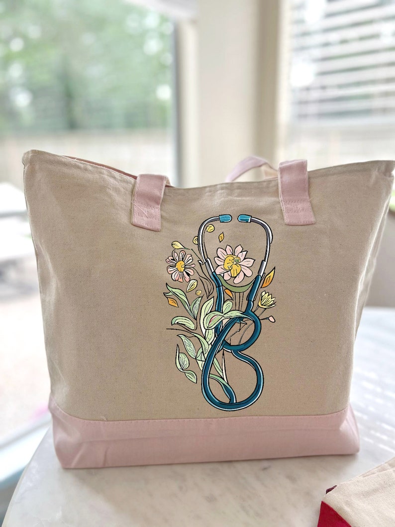 May include: A beige canvas tote bag with pink handles and a pink bottom. The bag has a floral design with a stethoscope in the center.