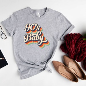 May include: Gray t-shirt with a retro-style graphic that says "90's Baby" in rainbow colors.