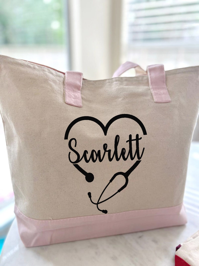 May include: A white canvas tote bag with pink handles and trim. The bag has a black heart design with a stethoscope and the name "Scarlett" inside the heart.