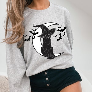 May include: Light gray sweatshirt featuring a black cat wearing a witch hat, sitting on a white crescent moon. Black bats are flying around the cat and moon. The sweatshirt is a comfortable, casual style.