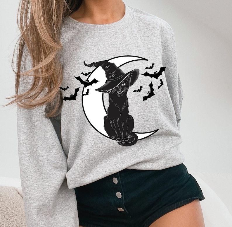 May include: Light gray sweatshirt featuring a black cat wearing a witch hat, sitting on a white crescent moon. Black bats are flying around the cat and moon. The sweatshirt is a comfortable, casual style.