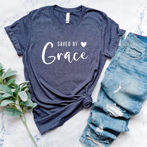 K&ouml;nnte beinhalten: Ein dunkelgraues T-Shirt mit dem Text "Saved by Grace" in wei&szlig;en Buchstaben mit einem kleinen wei&szlig;en Herz. Das Shirt ist an den &Auml;rmeln hochgekrempelt und liegt auf einer wei&szlig;en Marmorfl&auml;che.