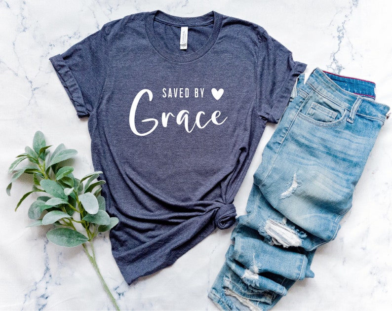 K&ouml;nnte beinhalten: Ein dunkelgraues T-Shirt mit dem Text "Saved by Grace" in wei&szlig;en Buchstaben mit einem kleinen wei&szlig;en Herz. Das Shirt ist an den &Auml;rmeln hochgekrempelt und liegt auf einer wei&szlig;en Marmorfl&auml;che.
