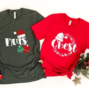 May include: Two Christmas-themed t-shirts. One is dark green with the word "nuts" and a Santa hat graphic, along with two ornaments. The other is red with the word "chest" inside a wreath and a Santa hat graphic.