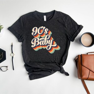 May include: A dark gray t-shirt with a retro-style graphic that says "90's Baby" in colorful, rainbow-like lettering.