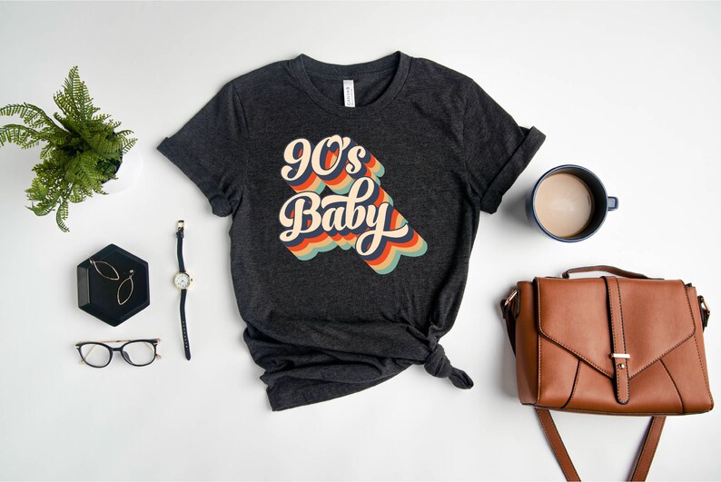 May include: A dark gray t-shirt with a retro-style graphic that says "90's Baby" in colorful, rainbow-like lettering.