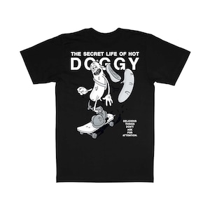 以下が含まれることがあります： スケートボードに乗ったホットドッグの漫画のキャラクターが描かれた黒いTシャツ。「THE SECRET LIFE OF HOT DOGGY」と「DELICIOUS THINGS DON'T ASK FOR ATTENTION」の文字がプリントされています。