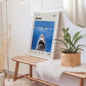Op de afbeelding: Een ingelijste poster van de film "Jaws", met een haai die uit de oceaan komt. De poster heeft een witte lijst en staat op een houten bank. Het ontwerp bevat het jaar 1975. Een decoratief item.