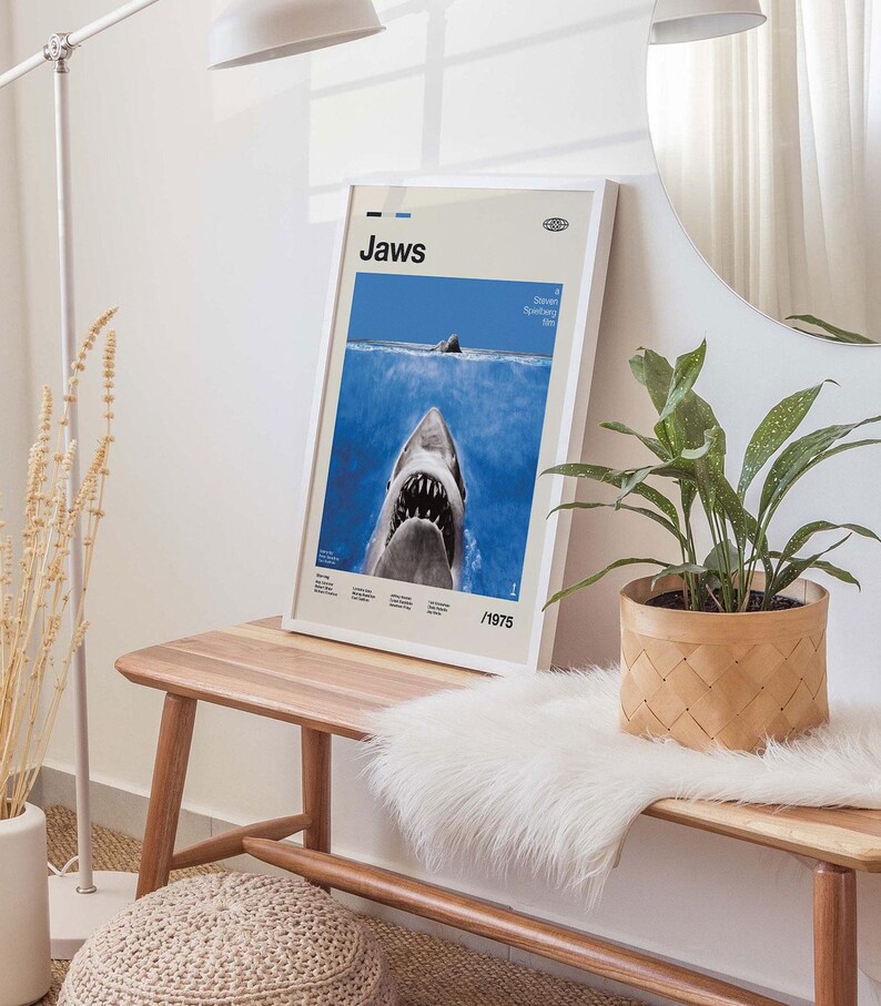 Op de afbeelding: Een ingelijste poster van de film "Jaws", met een haai die uit de oceaan komt. De poster heeft een witte lijst en staat op een houten bank. Het ontwerp bevat het jaar 1975. Een decoratief item.