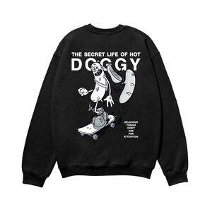 以下が含まれることがあります： 黒いスウェットシャツ。ホットドッグのキャラクターがスケートボードに乗ったグラフィックが特徴です。「THE SECRET LIFE OF HOT DOGGY」の文字が上にあります。グラフィックの下には「DELICIOUS THINGS DON'T ASK FOR ATTENTION」と書かれています。