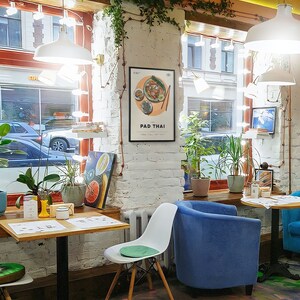 May include: A white brick wall with a framed poster that says "PAD THAI" and a picture of a plate of food. There is a blue armchair and a white chair with a green cushion. There is a wooden table with a yellow vase and a white cup on it.