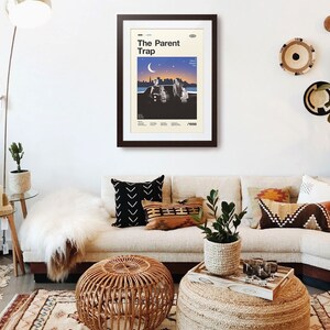 May include: A framed poster reading "The Parent Trap" in a living room. The poster has a retro design with a cityscape and figures under a crescent moon. The room features a beige sofa, decorative cushions, and a woven ottoman.
