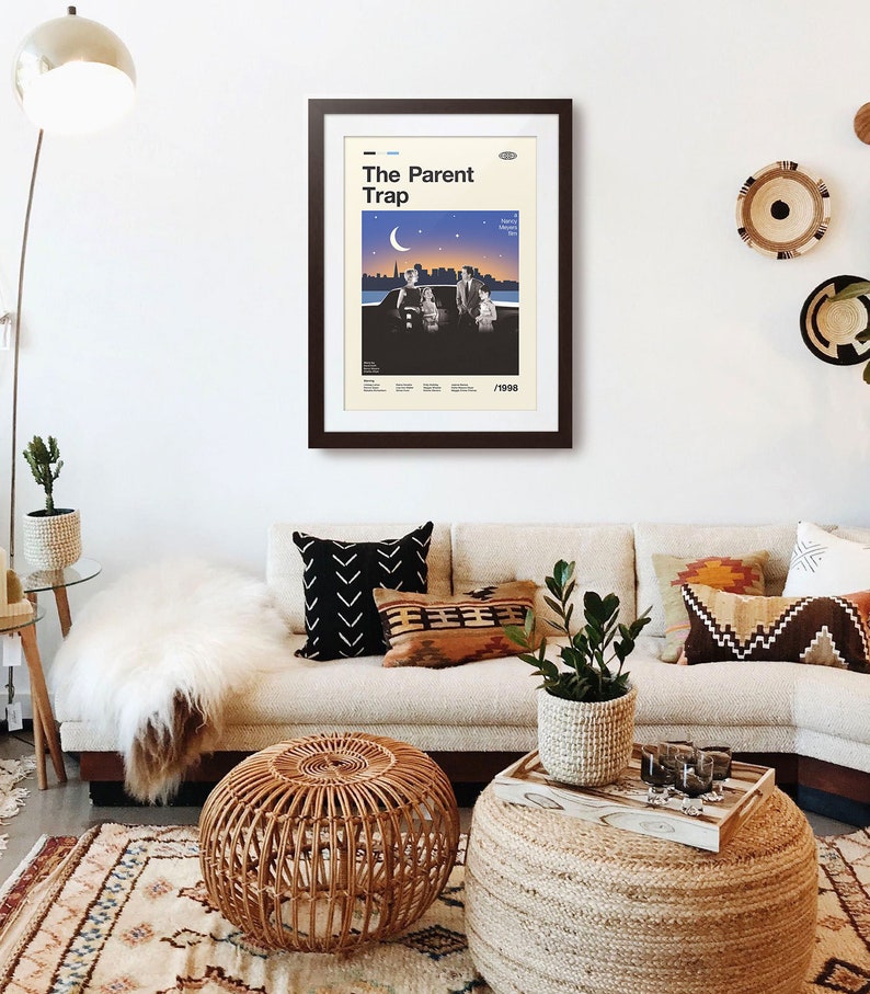 May include: A framed poster reading "The Parent Trap" in a living room. The poster has a retro design with a cityscape and figures under a crescent moon. The room features a beige sofa, decorative cushions, and a woven ottoman.