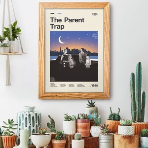 May include: A framed movie poster for "The Parent Trap" in a wooden frame. The poster shows a family scene with a cityscape and a crescent moon against a starry sky. The poster is on a white wall, surrounded by potted plants and succulents.