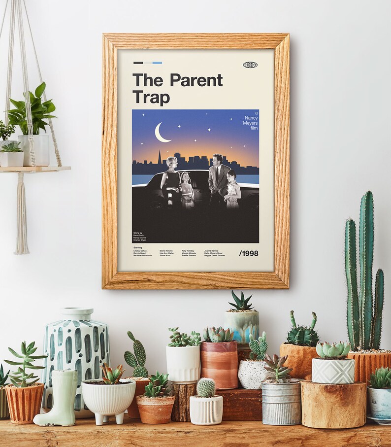 May include: A framed movie poster for "The Parent Trap" in a wooden frame. The poster shows a family scene with a cityscape and a crescent moon against a starry sky. The poster is on a white wall, surrounded by potted plants and succulents.