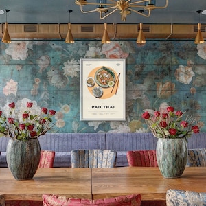 May include: A framed poster with a vintage illustration of Pad Thai, a popular Thai noodle dish. The poster is hanging on a wall with a floral pattern and is surrounded by a blue-grey wall.
