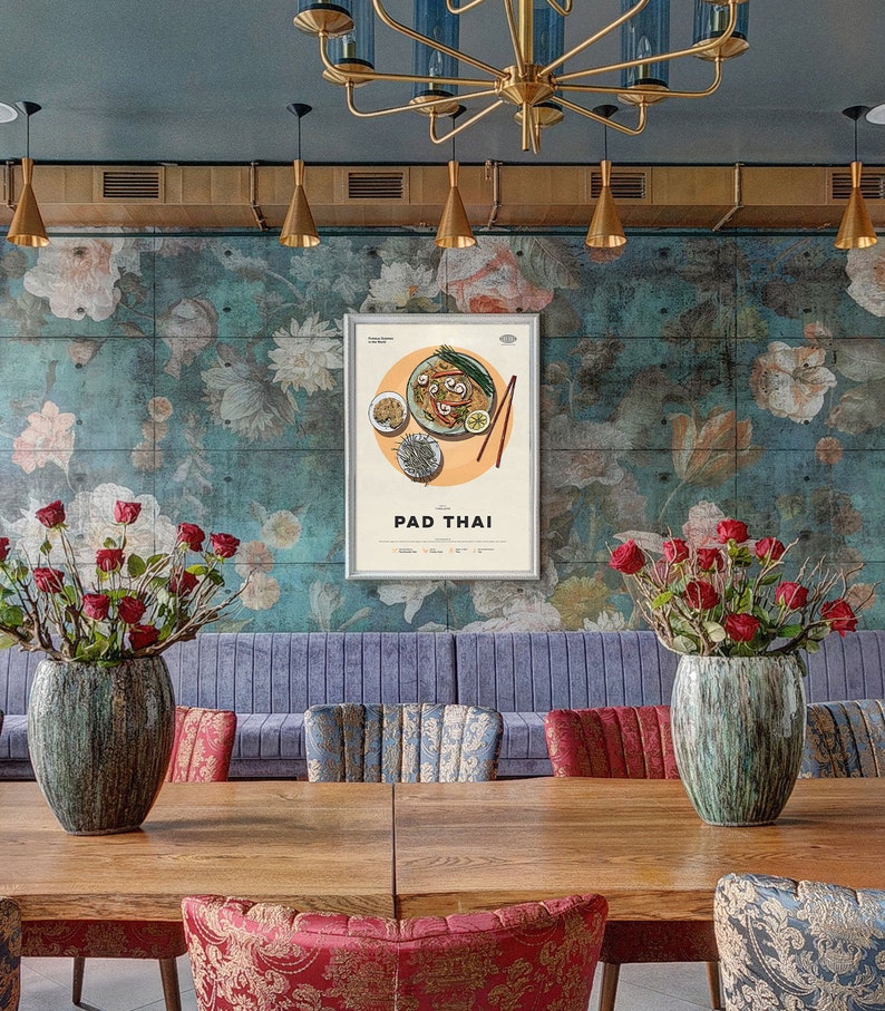 May include: A framed poster with a vintage illustration of Pad Thai, a popular Thai noodle dish. The poster is hanging on a wall with a floral pattern and is surrounded by a blue-grey wall.