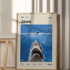 Op de afbeelding: Een ingelijste filmposter voor "Jaws". De poster toont een blauwe oceaansc&egrave;ne met de open kaken van een witte haai op de voorgrond. De titel "Jaws" staat bovenaan, met de naam van de regisseur en het jaar 1975.