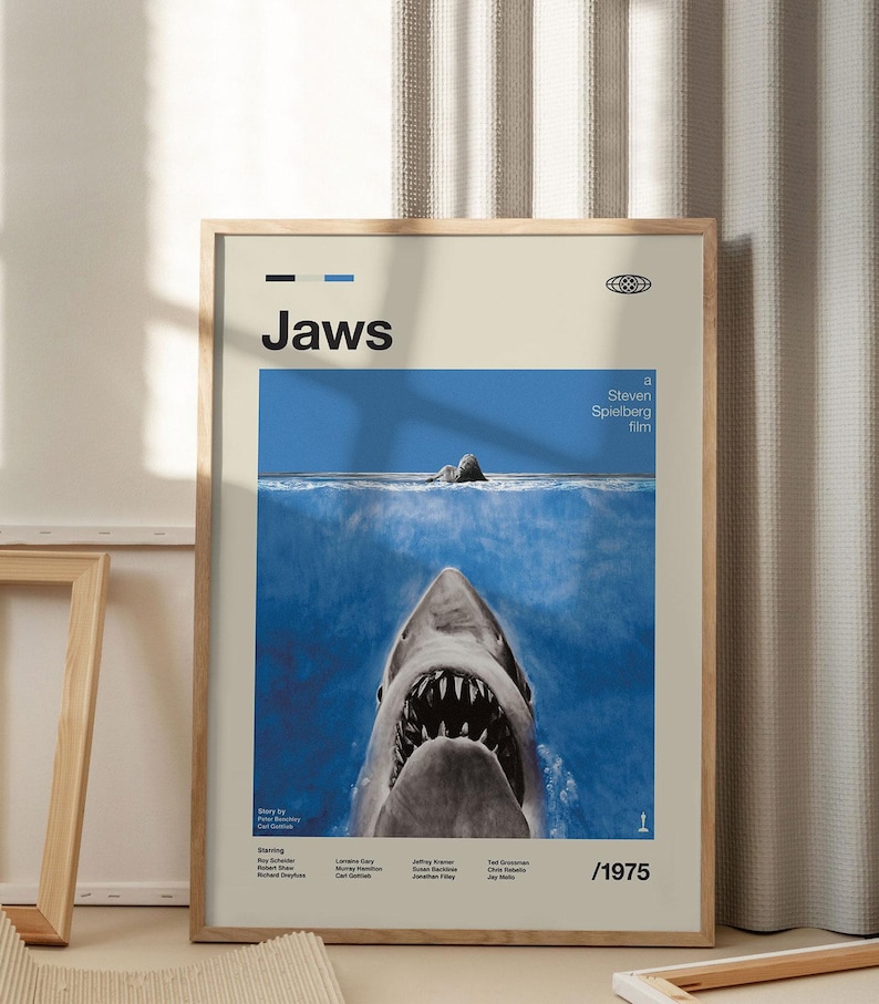Op de afbeelding: Een ingelijste filmposter voor "Jaws". De poster toont een blauwe oceaansc&egrave;ne met de open kaken van een witte haai op de voorgrond. De titel "Jaws" staat bovenaan, met de naam van de regisseur en het jaar 1975.