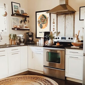 May include: A modern kitchen with white cabinets, a stainless steel oven and range, and a brown woven rug. A framed print with the text "Pad Thai" hangs on the wall above the stove.