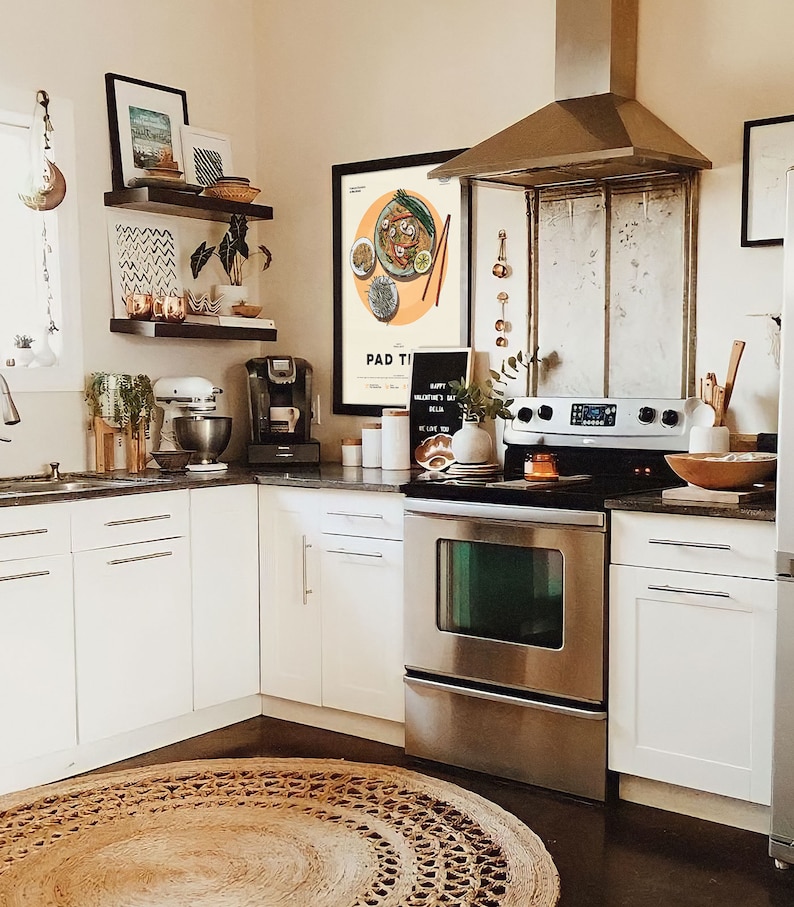May include: A modern kitchen with white cabinets, a stainless steel oven and range, and a brown woven rug. A framed print with the text "Pad Thai" hangs on the wall above the stove.