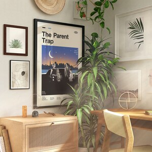 May include: An interior scene featuring framed art, a poster for "The Parent Trap" film, and a wooden cabinet. The room is adorned with plants, a woven wall hanging, and a wooden desk and chair. The colour palette is neutral with natural wood tones.