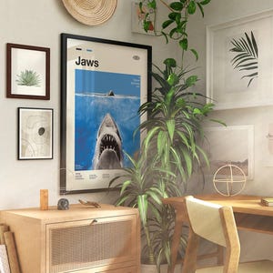 Op de afbeelding: Een kamer met ingelijste kunstwerken en een houten bureau. Het middelpunt is een ingelijste "Jaws" filmposter met een haai. Andere ingelijste kunstwerken zijn een abstracte print, een botanische print en een palmbladprint.