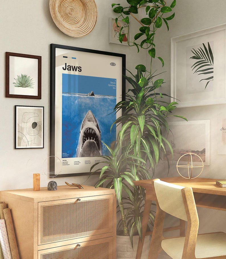 Op de afbeelding: Een kamer met ingelijste kunstwerken en een houten bureau. Het middelpunt is een ingelijste "Jaws" filmposter met een haai. Andere ingelijste kunstwerken zijn een abstracte print, een botanische print en een palmbladprint.