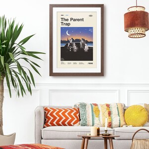 May include: A framed poster with the title "The Parent Trap" in a retro style. The poster, in a brown frame, shows a family scene under a night sky, with the year 1998 at the bottom. It hangs above a grey sofa with colourful cushions, creating a cosy living room setting.