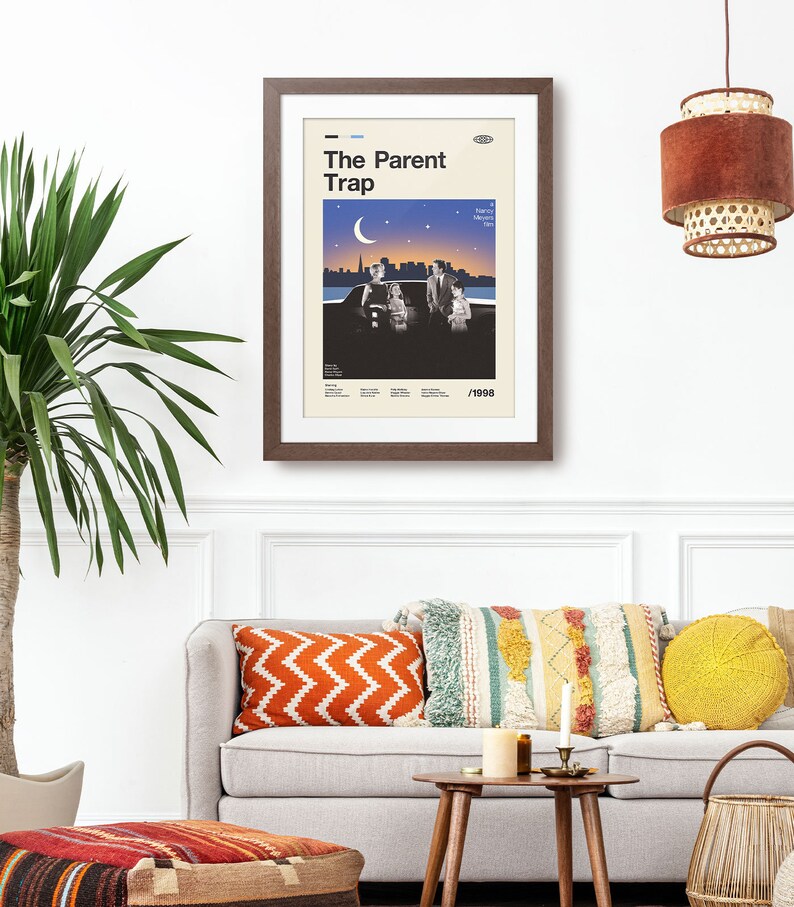 May include: A framed poster with the title "The Parent Trap" in a retro style. The poster, in a brown frame, shows a family scene under a night sky, with the year 1998 at the bottom. It hangs above a grey sofa with colourful cushions, creating a cosy living room setting.