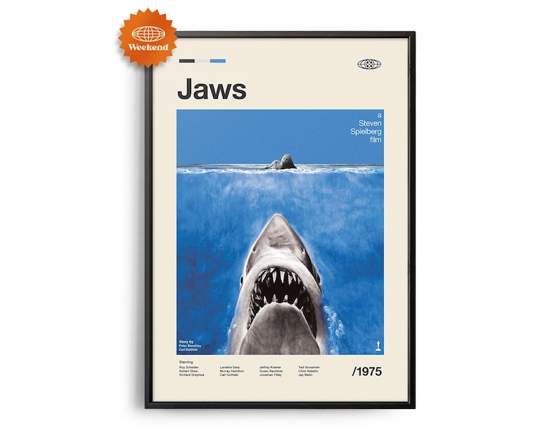 Op de afbeelding: Ingelijste filmposter voor de film "Jaws" met een zwart frame. De poster toont de open bek van een haai in het water, met de titel "Jaws" in grote witte letters. De poster bevat ook het jaar 1975.