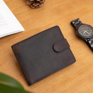 Puede incluir: Una cartera plegable de cuero marrón oscuro con cierre de broche y la palabra Dad grabada en la parte frontal, junto a un reloj de eslabones de metal oscuro con esfera negra, un libro abierto y dos pequeñas piñas de pino.
